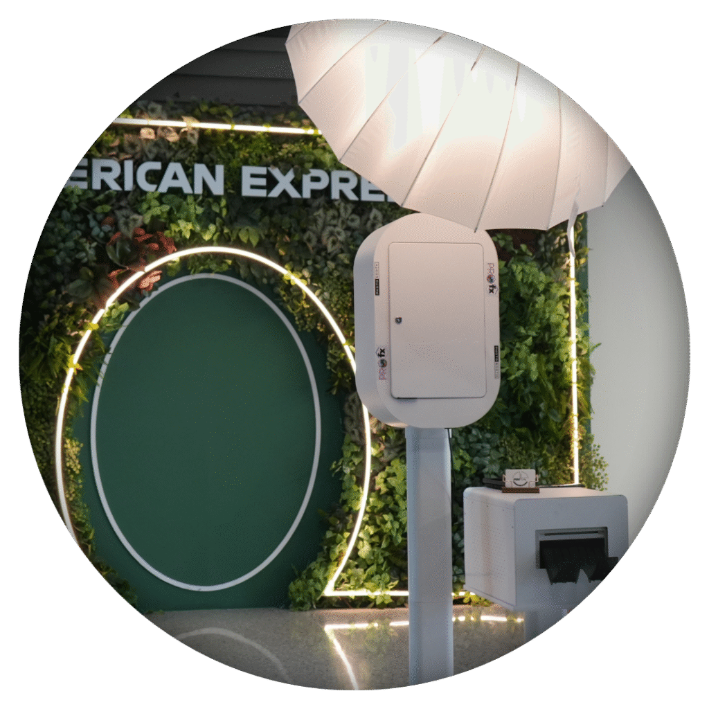 Stylish white photo booth station with studio lighting in front of an elegant greenery wall branded with American Express signage.
