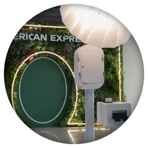 Stylish white photo booth station with studio lighting in front of an elegant greenery wall branded with American Express signage.