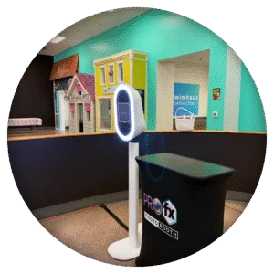 Modern digital photo booth setup with sleek white stand and branded Pro FX display in a colorful indoor setting.